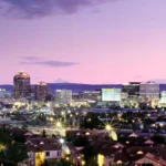 A cityscape at dusk, with illuminated buildings and residential areas under a purple and pink sky.