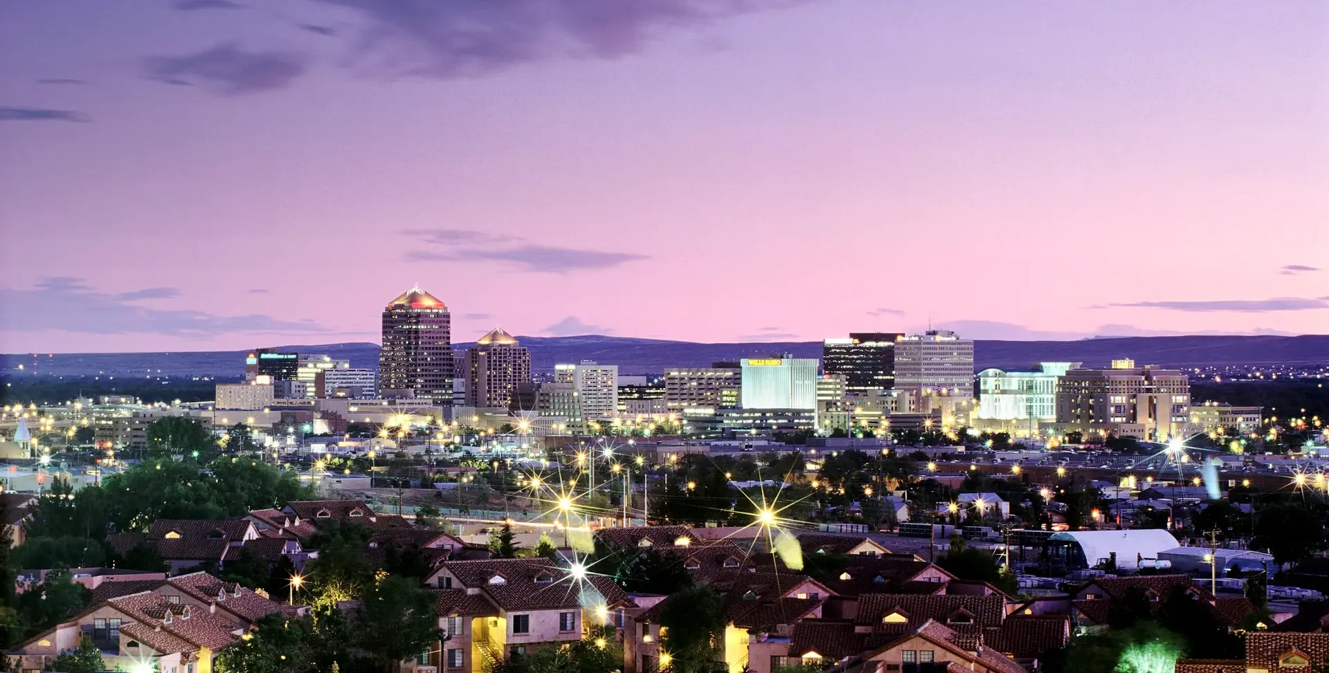 A cityscape at dusk, with illuminated buildings and residential areas under a purple and pink sky.